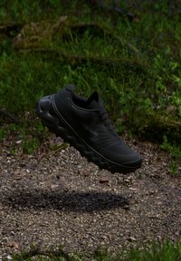 Chaussure de sport noire avec un dessus texturé, un design léger et un profil de semelle épaisse et ondulée, flottant au-dessus d'une surface en gravier.