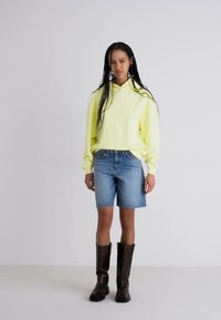 Felpa gialla con cappuccio, pantaloni corti in denim blu e stivali marroni fino al ginocchio; il modello è in piedi contro uno sfondo bianco uniforme.