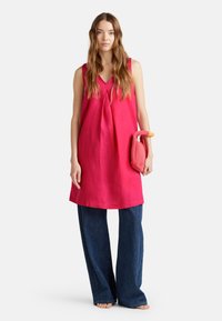 Fuchsia farbenes Leinen-Tunikakleid ohne Ärmel mit V-Ausschnitt, kombiniert mit weiten, dunklen Jeans. Das Model hält eine kleine, strukturiert gestaltete rosa Tasche.