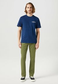 Marineblauwe katoenen t-shirt met korte mouwen en een klein wit logo op de borst, gecombineerd met olijfgroene slim-fit broeken en zwarte sneakers.