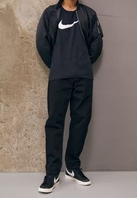 Svart bomberjacka över en svart t-shirt med en vit Nike-logotyp, tillsammans med svarta byxor och svarta sneakers med vita accenter.