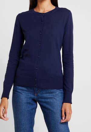 Woman wearing a navy blue buttoned cardigan with long sleeves and blue jeans, standing against a plain background.