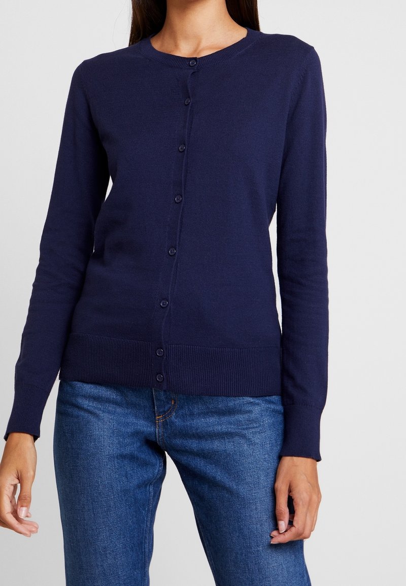 Femme portant un cardigan bleu marine boutonné à manches longues et un jean bleu, posant devant un fond uni.