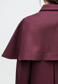 Burgundy coat with a structured cape design, featuring a smooth texture and collar, shown from a back-angle view.