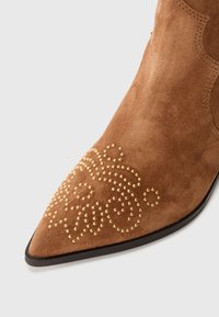 Botín de ante marrón de punta afilada que presenta un intrincado detalle de tachuelas doradas y una suela negra elegante. Textura suave con acentos decorativos.