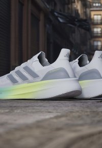 Paire de chaussures de course Adidas Ultraboost blanches avec des bandes grises et des semelles intermédiaires à dégradé vert clair reposant sur un pavé en pierre.