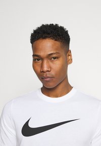 Hvid langærmet T-shirt lavet af bomuld med et fremtrædende sort Nike Swoosh-logo på tværs af brystet. Rent, minimalistisk design.