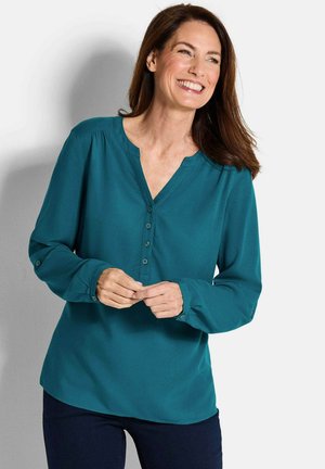 Tüpfelbluse mit V-Ausschnitt, Knopfleiste und langen Ärmeln. Glatter Stoff und dezente Falten an den Schulternähten. Kombinieren Sie sie mit dunklen Jeans.