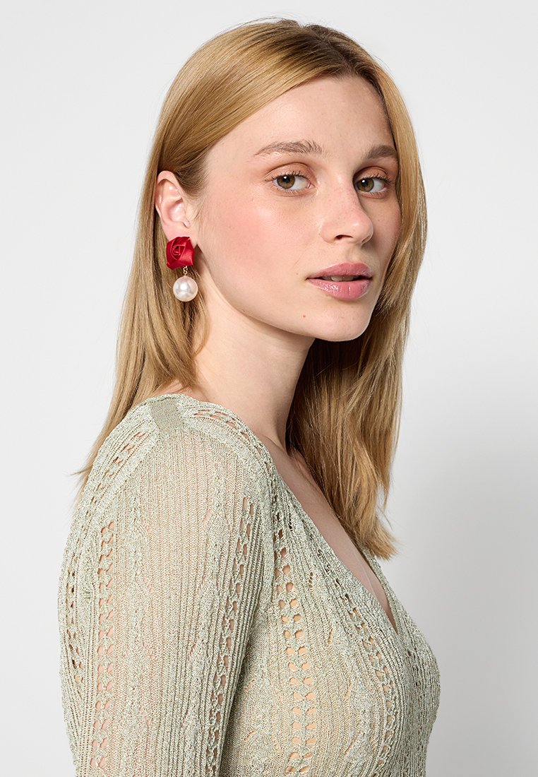 Orecchini a forma di rosa rossa con goccia di perla, hardware in oro e design texture. Il modello indossa un top lavorato a maglia verde chiaro.