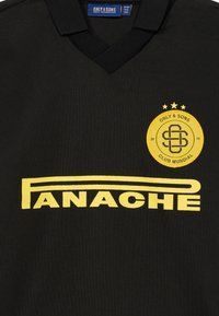 Schwarzes Polohemd mit V-Ausschnitt. Es hat gelb bedruckten Text "FANACHE" und ein rundes Logo mit drei Sternen in Gelb.