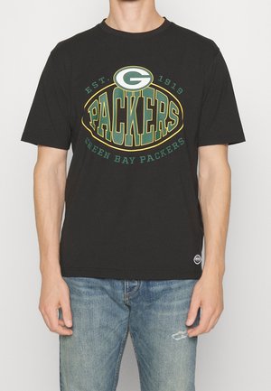 Personne portant un t-shirt noir des Green Bay Packers avec le logo et le texte, accompagné d'un jean bleu clair déchiré.