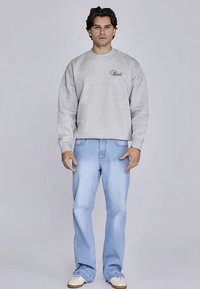 Gray sweatshirt with embroidered logo, paired with light blue jeans. Model wears cream sneakers; stands against a plain light background.