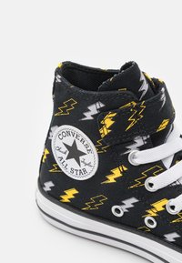 Zapatilla alta negra con un patrón de rayos en amarillo y gris, cordones blancos y un logo circular de "Converse All Star". Suela de goma.