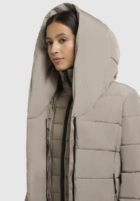 Veste matelassée beige avec une grande capuche, un design matelassé, une fermeture éclair à l'avant et des accents de fermeture éclair noirs. Texture lisse et douce.