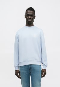 Svetlomodrá mikina z bavlnenej zmesi, s okrúhlym výstrihom, dlhými rukávmi a malým bielym logom na hrudi. Kombinovaná s džínsami.
