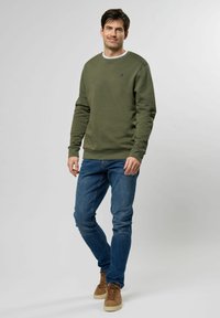 Olajzöld pulóver bordás nyakkivágással és mandzsettákkal, kék farmernadrággal és világosbarna velúr sportcipővel párosítva. Egyszerű dizájn, laza fazon.