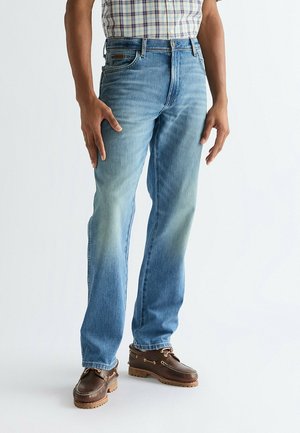 Mężczyzna ubrany w jasnoniebieskie jeansy o prostych nogawkach, brązowe skórzane buty żeglarskie oraz krótki rękaw koszuli w kratę, stojący na jednolitym tle.