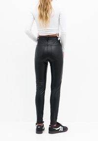 Zwarte nep leren leggings met een hoge taille en een aansluitend ontwerp. Bevat achterzakken en een gladde, glanzende afwerking. Draagt met een witte top en sneakers.