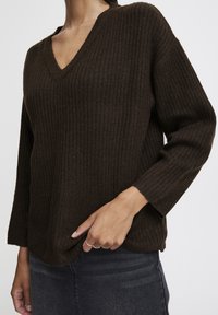 Jersey de punto marrón con cuello en V, mangas largas y un corte relajado, que presenta una textura sutil y bordes rematados.