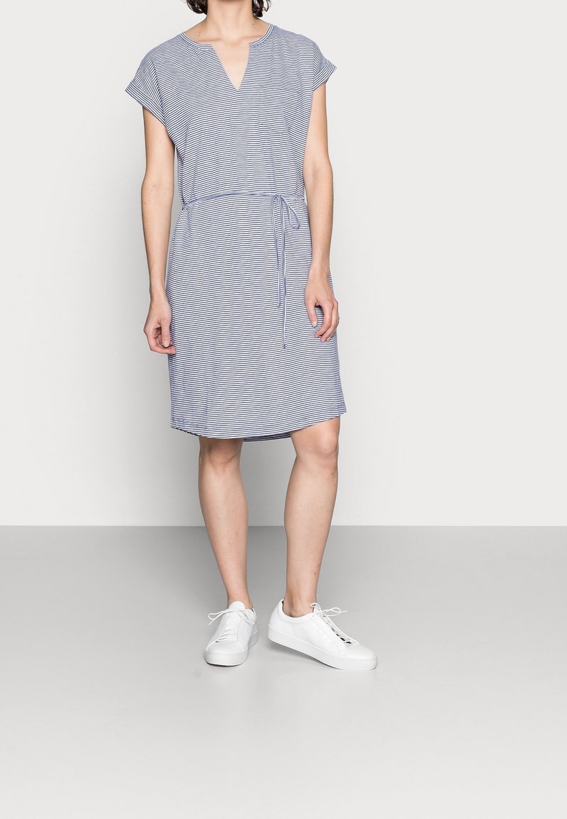 Robe en coton à rayures bleu et blanc, avec un col en V, des manches courtes, un détail noué à la taille, et un ourlet droit. Associée à des baskets blanches.