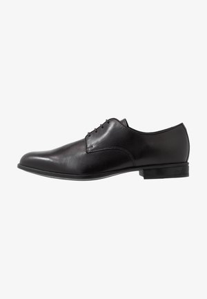 Chaussure habillée en cuir noir à finition lisse, bout arrondi, design à cinq œillets avec lacets, et coutures minimalistes. Talon bas en caoutchouc.