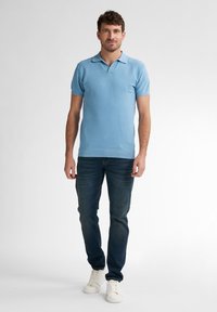 Světle modré polo tričko s krátkým rukávem a texturovaným vzorem, v kombinaci s tmavě modrými džínami a bílými teniskami. Model stojí v neutrálním prostředí.
