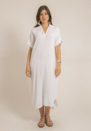 Femme portant une robe midi blanche texturée avec manches courtes, col, fente latérale, grandes boucles d'oreilles florales, un bracelet et des sandales cloutées.
