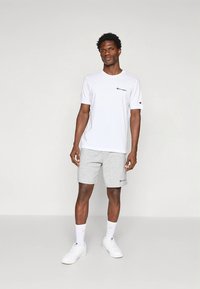 White cotton t-shirt with a small black logo on the chest, paired with light gray shorts. Model wears white sneakers and socks.