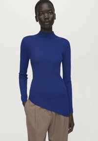 Modri rebrasti pulover z visokim ovratnikom in asimetričnim robom, dolgimi rokavi in oprijeto zasnovo, kombiniran z bež hlačami z visokim pasom.