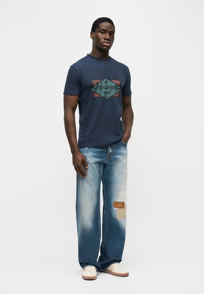 Άνδρας με μπλε ναυτικό μπλουζάκι με σχέδιο, φαρδιά ξεθωριασμένα μπλε τζιν με μπαλώματα και λευκά αθλητικά παπούτσια, στέκεται με το ένα χέρι στην τσέπη.