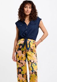 Chemise bleu marine à boutons avec manches retroussées et deux poches ; pantalon jaune à motif floral avec accents roses, bleus et verts ; détail de taille nouée.