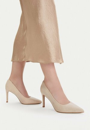 Frau trägt einen beigen Satin-Midirock und beigefarbene spitze High Heels mit schlanken Stiletto-Absätzen vor weißem Hintergrund.