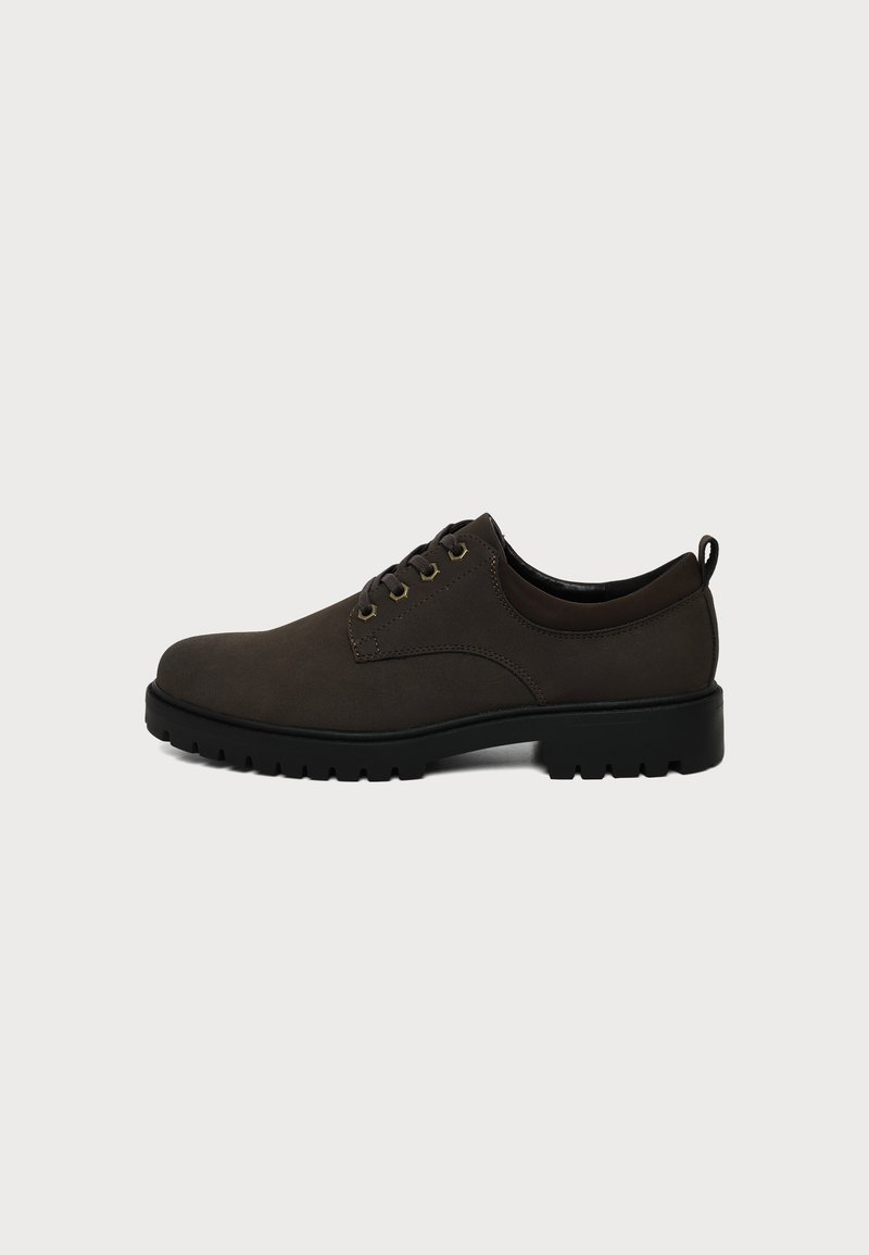 Chaussure en cuir marron foncé à lacets avec semelle épaisse en caoutchouc noir et languette à l’arrière, vue de côté sur un fond uni.