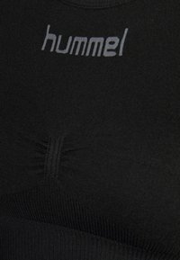 Svart sports-bh med en glatt, elastisk tekstur. Har samlet detalj foran og "hummel"-logoen i grått på den øverste delen.