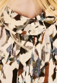 Acercamiento de un cuello de blusa con dos botones dorados y un patrón abstracto en tonos marrón, negro y beige, enmarcado por mechones de cabello rubio.