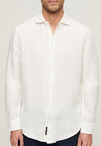 Camisa blanca de lino con botones, de corte relajado y mangas largas, que presenta una textura sutil y una etiqueta en el dobladillo del lado izquierdo.