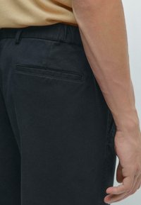 Pantalons en coton noir avec une texture lisse, taille élastique et une poche arrière unique. Présenté de côté, montrant le placement des mains.