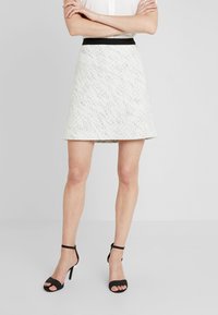 Jupe a-line blanche texturée avec une taille noire, assortie de sandales à talons noires. Le tissu présente un motif à pois subtil.