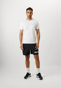 Hvid kortærmet t-shirt med logo på brystet, kombineret med sorte shorts med 'NIKE' i stor tekst. Sorte sneakers og hvide sokker.