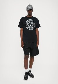 Svart t-skjorte med korte ermer og en hvit Versace Jeans Couture-logo, kombinert med svarte shorts og svarte joggesko, samt en grå cap.