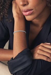 Bracelet en argent avec de petites pierres claires et rondes, porté sur un bras bronzé à la finition texturée. Chemise en tissu noir visible en arrière-plan.