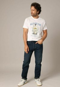 T-shirt en coton blanc avec logo graphique bleu, associé à un jean bleu foncé. Le modèle porte des baskets blanches avec des accents bleus. Tenue simple et décontractée.