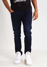 Tmavě modré džíny s úzkým střihem, vyrobené z denimu, s jemnou strukturou a pěti kapsami, v kombinaci s bílými sportovními botami.