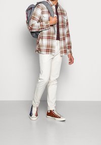 Geruite shirt in bruin, wit en beige; bordeaux T-shirt eronder; crème broek; bruin en blauwe sneakers; grijze rugzak met schouderbanden.