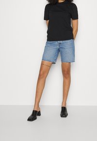 Camisa negra de mangas abullonadas combinada con shorts de mezclilla azul claro, con dobladillo deshilachado, y mule de tacón bloque negro. Un atuendo simple y casual.
