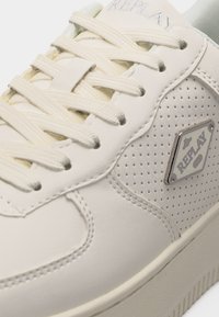 Baskets blanches perforées avec lacets crème et logo en métal de la marque Replay sur le côté, vue rapprochée de la tige et de la semelle.