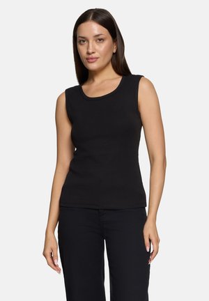 Femme debout portant un haut noir sans manches et un pantalon noir sur un fond clair uni.