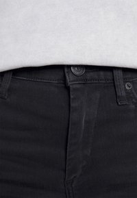 Gros plan sur la taille d'un jean en denim noir avec bouton et passants pour ceinture, associé à une chemise claire rentrée à l'intérieur.