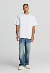 Biała bawełniana koszulka z krótkim rękawem w połączeniu z jasnoniebieskimi dżinsami. Model stoi z neutralnym wyrazem twarzy, nosząc białe trampki.