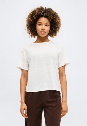 Νέα γυναίκα με σγουρά μαλλιά φοράει λευκή κοντομάνικη μπλούζα και καφέ παντελόνια, στέκεται μπροστά σε ένα απλό ανοιχτό φόντο.